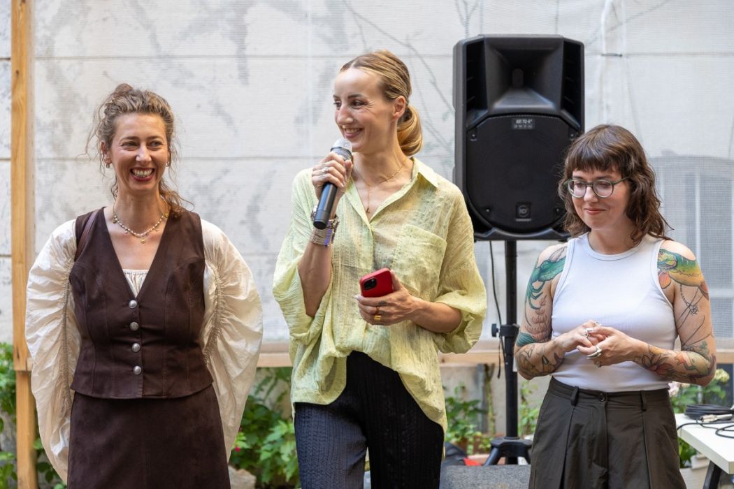 Three women stand side by side in a bright indoor space. The smiling woman in the centre speaks into a microphone and holds a red phone in her other hand, while the women beside her smile. A large loudspeaker is visible in the background.