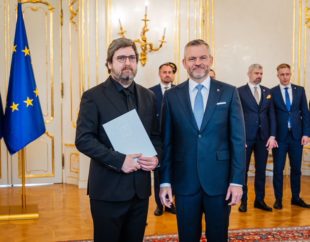 Two people in formal attire at an official ceremony in a ceremonial room, a woman holding a document, with EU and Slovak flags in the background.