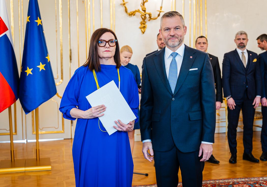 Two people in formal attire at an official ceremony in a ceremonial room, a woman holding a document, with EU and Slovak flags in the background.