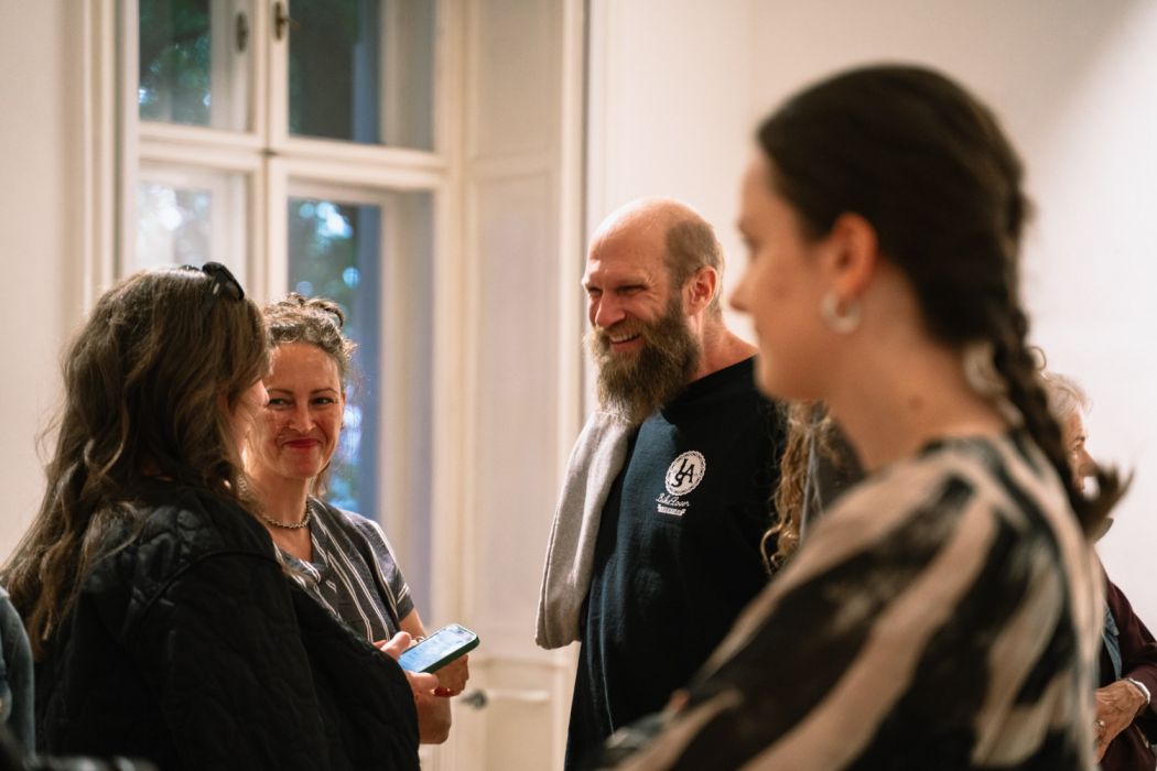 Fotografia skupiny návštevníkov vo svetlom interiéri galérie pri veľkých oknách. Vľavo sa rozprávajú dve ženy, jedna drží telefón; v strede sa usmieva bradatý muž a v popredí vpravo je rozostrená žena s vrkočom.