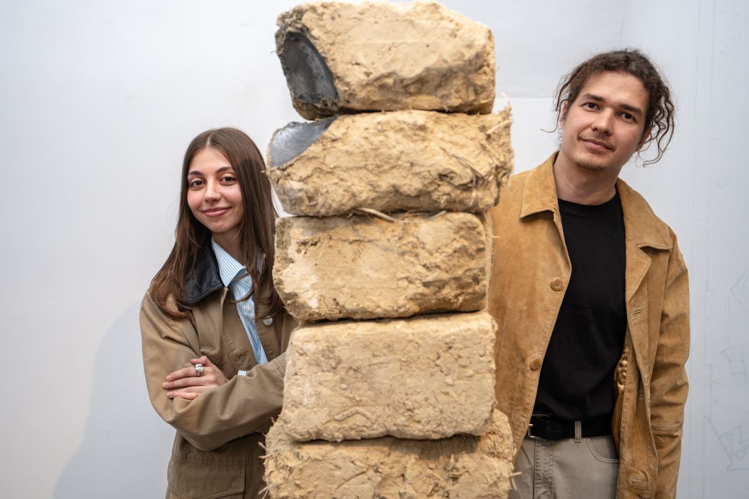 Dvaja ľudia stoja pri stĺpovej soche z hrubých materiálov, ktorá je zložená z vrstvených blokov s kovovými prvkami.