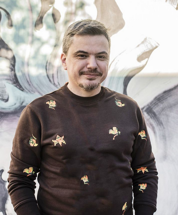 Portrét muža v tmavom svetri s drobným vzorom, stojí pred abstraktnou maľbou.