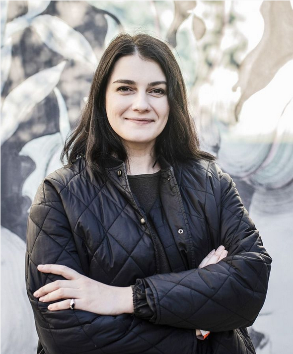 Portrét ženy v čiernej bunde so skríženými rukami, stojí pred abstraktnou maľbou.