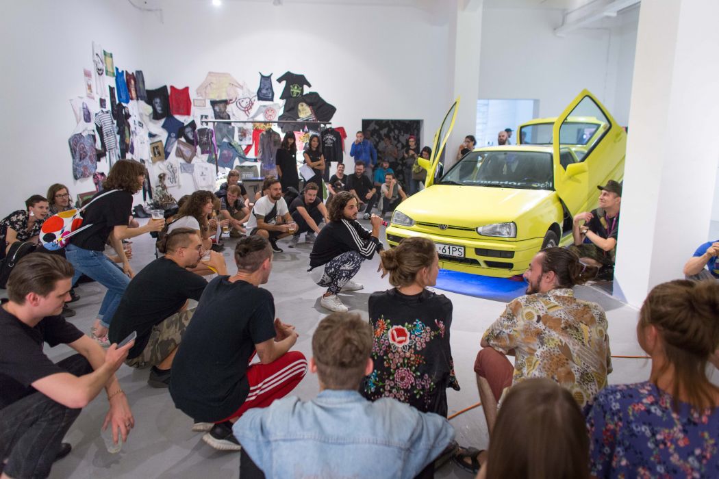A group of people sit and squat in a gallery space around a yellow car with open doors during a performative or discussion-based event. T-shirts and graphic works are displayed on the walls, and the audience informally gathers around the central action.